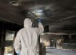 tecnico in tuta protettiva ispeziona una griglia di ventilazione annerita dalla fuliggine durante la bonifica delle condotte dopo un incendio
