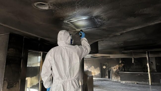 Bonifica condotte dopo incendio: sopralluogo su griglia annerita tecnico in tuta protettiva ispeziona una griglia di ventilazione annerita dalla fuliggine durante la bonifica delle condotte dopo un incendio