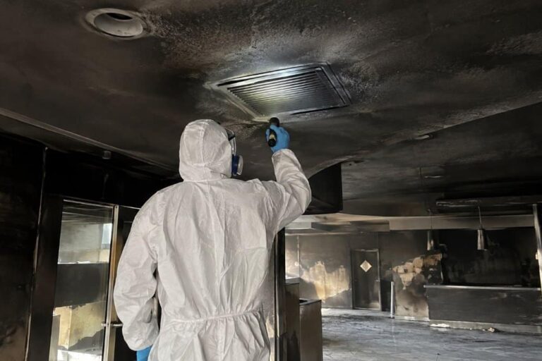 tecnico in tuta protettiva ispeziona una griglia di ventilazione annerita dalla fuliggine durante la bonifica delle condotte dopo un incendio