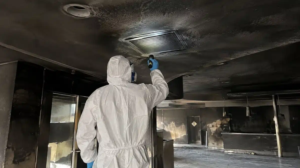 tecnico con tuta protettiva e maschera ispeziona una condotta aeraulica coperta di fuliggine in un ambiente danneggiato da un incendio