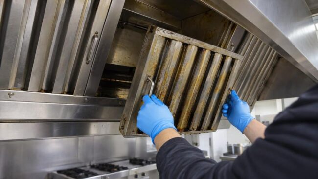 tecnico con guanti in nitrile rimuove un filtro a labirinto coperto di grasso dalla cappa di una cucina professionale durante la pulizia dei canali grassi